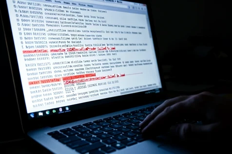 A close-up of a laptop terminal window filled with monospaced log output, one line highlighted in red reading "onnxruntime CUDAExecutionProvider failed to load", with surrounding white and grey log lines slightly out of focus. A hand rests near the trackpad. Setting is a dim home workspace at night. The screen is the only light source, casting a cool blue-white glow upward onto the keyboard and the edge of the hand, hard-edged and contrasty against the dark room. Tense, debugging-late mood.