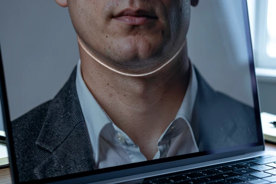 A close-up macro shot of a single AI-generated headshot displayed on a laptop screen, zoomed to 200% so a thin lighting seam at the subject's jawline and neck is visible as a faint horizontal band where the warmer face skin tone meets the cooler neck and shirt collar. The subject is a person in their thirties wearing a charcoal blazer over a crisp white shirt, set against a soft grey studio background. The laptop sits on a wooden desk, a paper notebook and a pen in the corner of the frame. Cool office daylight from the right, low diffuse fill from the left, slight reflection on the screen surface. Documentary, editorial photography style, calm muted atmosphere.