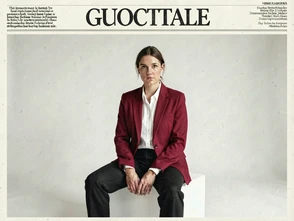 A person in bold editorial fashion — a structured crimson blazer over a white shirt — sits on a minimalist white cube against a clean studio backdrop, gazing directly at the camera with a composed, confident expression, the composition leaving generous space at the top for a masthead, soft diffused overhead lighting creating even skin tones and subtle shadow definition, atmosphere polished and aspirational. Editorial illustration in the visual language of newsprint broadsheet, evoking 1980s ink and serif columns on cream paper. Palette: #F1EBDB ground, #1B1816 ink, #8B0000 primary highlight, #2A2A26 supporting hue. Soft printed daylight with paper grain. Style: newsprint broadsheet. Mood: solemn.