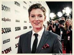 A person poses for a close-up portrait at a film premiere step-and-repeat banner, dressed in a tailored suit with a silk pocket square, smiling confidently at the camera, the branded backdrop covered in repeating studio logos, bright white press-line lights from directly in front creating a clean, even exposure on the face, shallow depth of field blurring the background crowd, atmosphere glamorous and high-profile. Editorial illustration in the visual language of newsprint broadsheet, evoking 1980s ink and serif columns on cream paper. Palette: #F1EBDB ground, #1B1816 ink, #8B0000 primary highlight, #2A2A26 supporting hue. Soft printed daylight with paper grain. Style: newsprint broadsheet. Mood: solemn.