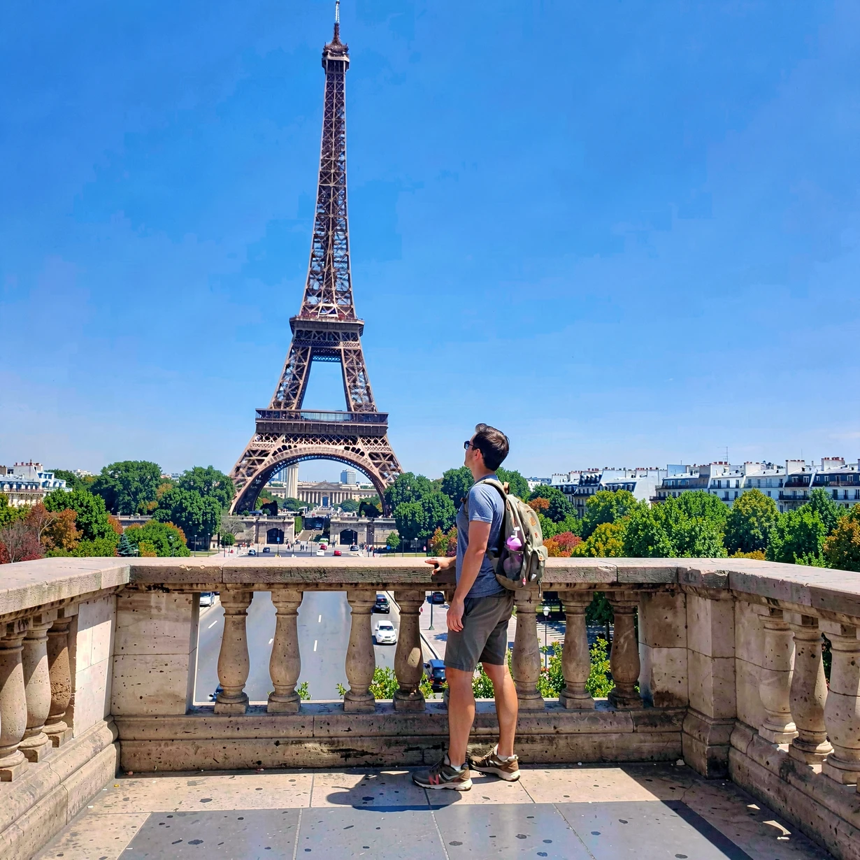 A solo traveler standing on a stone viewpoint platform overlooking the Eiffel Tower in Paris on a clear blue-sky afternoon, casual travel clothing and a small backpack, bright midday sunlight from above casting short crisp shadows, vibrant city colors, wide-angle travel photography composition with the landmark prominent in the background.