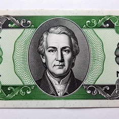 A close-up of a stylized banknote design featuring an engraved portrait in the center oval, surrounded by fine-line guilloche patterns and ornate borders in green and black ink, flat overhead lighting on a white surface, sharp macro detail revealing every engraved line, official currency illustration aesthetic.