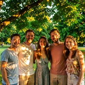 A group of six friends standing close together outdoors in a park on a sunny afternoon, all smiling naturally at the camera, dappled sunlight filtering through tree canopy above creating warm golden patches on their faces, casual summer clothing, vibrant green foliage background, candid lifestyle photography feel.