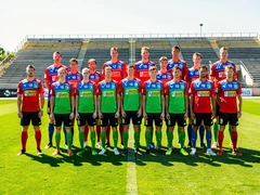 A group of athletes in matching team jerseys lined up in two rows on a green grass field for an official team photograph, bright midday sunlight from above casting short shadows, stadium bleachers visible in the background, sharp focus across all faces, vibrant team colors, clean sports photography composition.