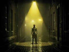 A lone figure in a long trench coat standing at the end of a fog-filled alley at night, dramatic single overhead streetlamp casting a hard cone of yellow light downward, deep shadows on either side, rain-wet cobblestones reflecting the light, moody cinematic noir atmosphere with high contrast and desaturated tones.